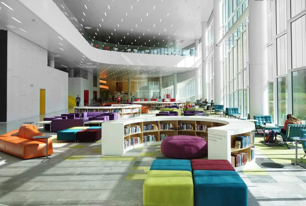 Modern library interior with floor-to-ceiling windows, colorful seating, and curved bookshelves under a high ceiling with recessed lighting. People are seen studying and reading in the bright, open space filled with natural light.