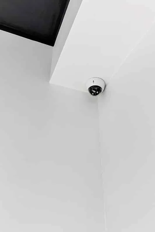 A dome security camera is mounted in the upper corner of a white ceiling, positioned to monitor the surrounding area. The minimal, clean setting emphasizes surveillance coverage in a modern interior space.