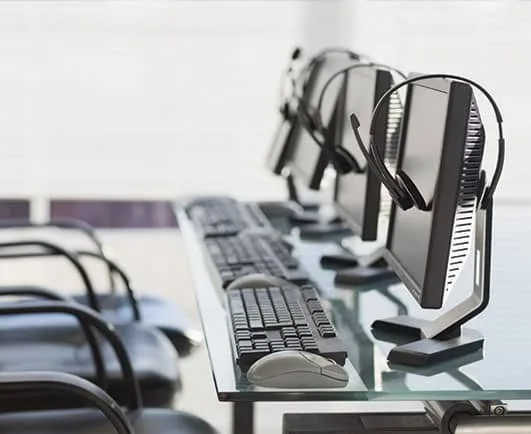 A row of computer monitors with headsets rests on a glass desk, each paired with a keyboard and mouse. Empty chairs line the setup, suggesting a call center, training room, or security operations workspace awaiting staff.