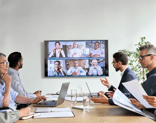 A group of professionals sits around a conference table engaged in a hybrid meeting, with several colleagues joining remotely via video call displayed on a wall-mounted screen. Laptops, documents, and glasses of water are spread across the table, highlighting a collaborative and modern work environment.