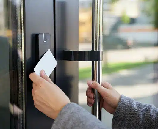 A person wearing a gray coat holds a key card up to an electronic access reader while gripping a glass door handle. The scene suggests secure entry into a building, emphasizing access control and physical security measures.