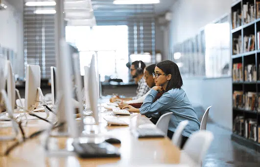 People work at desktop computers in a bright, modern library or computer lab. A woman in the foreground wears glasses and focuses on her screen, with shelves of books visible in the background.