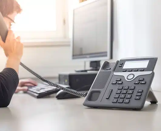 A close-up of a modern office desk phone in the foreground, with a person in the background talking on the receiver while typing at a computer. The scene conveys workplace communication and multitasking in a professional setting.