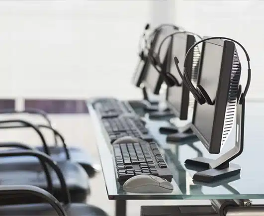 A row of computer monitors with headsets rests on a glass desk, each paired with a keyboard and mouse. Empty chairs line the setup, suggesting a call center, training room, or security operations workspace awaiting staff.