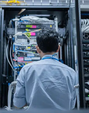 A network engineer seen from behind works inside an open server rack arranging color-tagged cables and devices. The tidy datacenter scene emphasizes precise setup and maintenance of critical network systems.