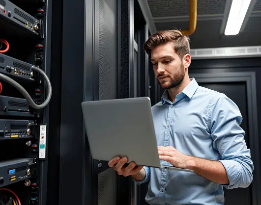 A man in a light blue dress shirt works on a laptop inside a server room, focused on monitoring or configuring the equipment. He stands next to server racks with visible cables and network hardware.