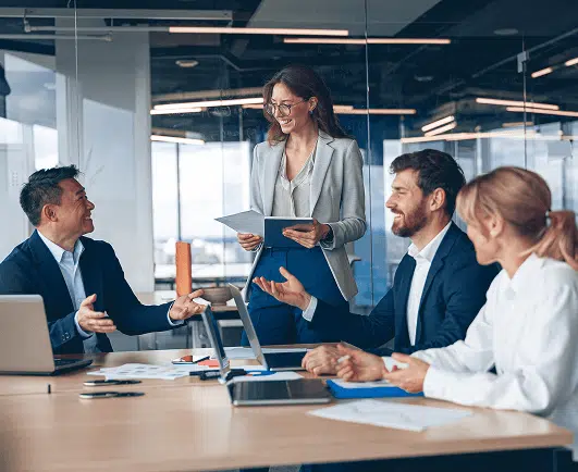A group of business professionals in a modern office shares a lighthearted discussion during a meeting, with one woman standing and holding documents while others sit around a table with laptops and papers. The atmosphere appears collaborative and upbeat.
