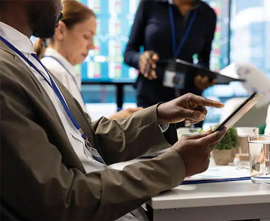A man in business attire uses a digital tablet during a meeting, while two colleagues, one seated and one standing, engage with documents in the background. Large digital stock market displays are visible through the window, suggesting a financial or data-driven work environment.