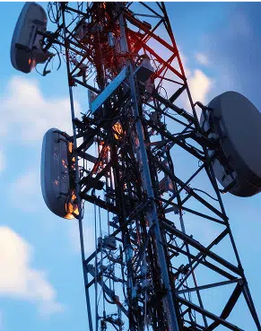 A close-up of a steel cell tower bristles with panel antennas and cables silhouetted against a soft blue evening sky. Warm red signal lights dot the structure emphasizing its role in wireless communication.