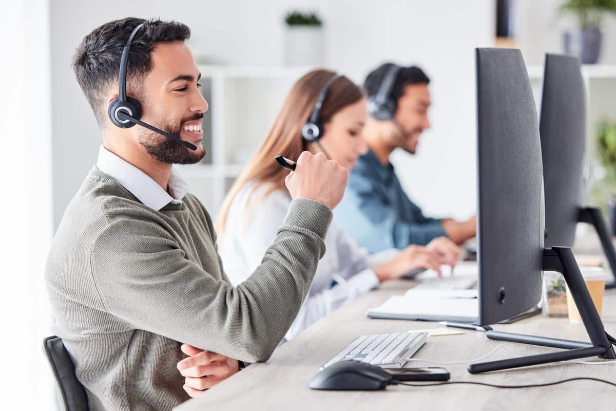 A cheerful man wearing a headset sits at a desk in a call center, holding a pen and looking at his computer monitor. Two other team members with headsets are working at computers in the background, creating a professional and collaborative environment.