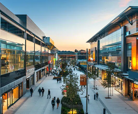 An open-air shopping center at sunset features modern architecture, glass storefronts, and well-lit walkways lined with small trees. Shoppers casually stroll between stores, creating a lively yet relaxed atmosphere.
