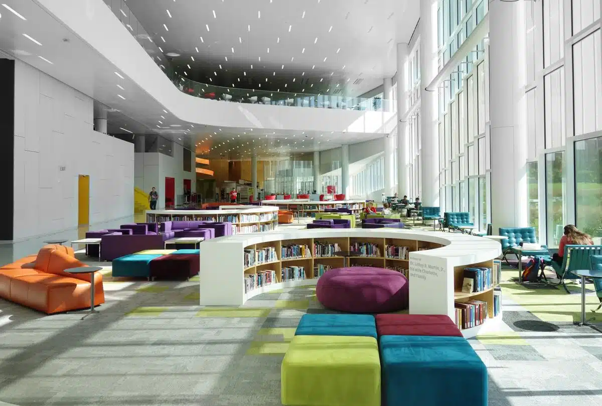 Modern library interior with floor-to-ceiling windows, colorful seating, and curved bookshelves under a high ceiling with recessed lighting. People are seen studying and reading in the bright, open space filled with natural light.