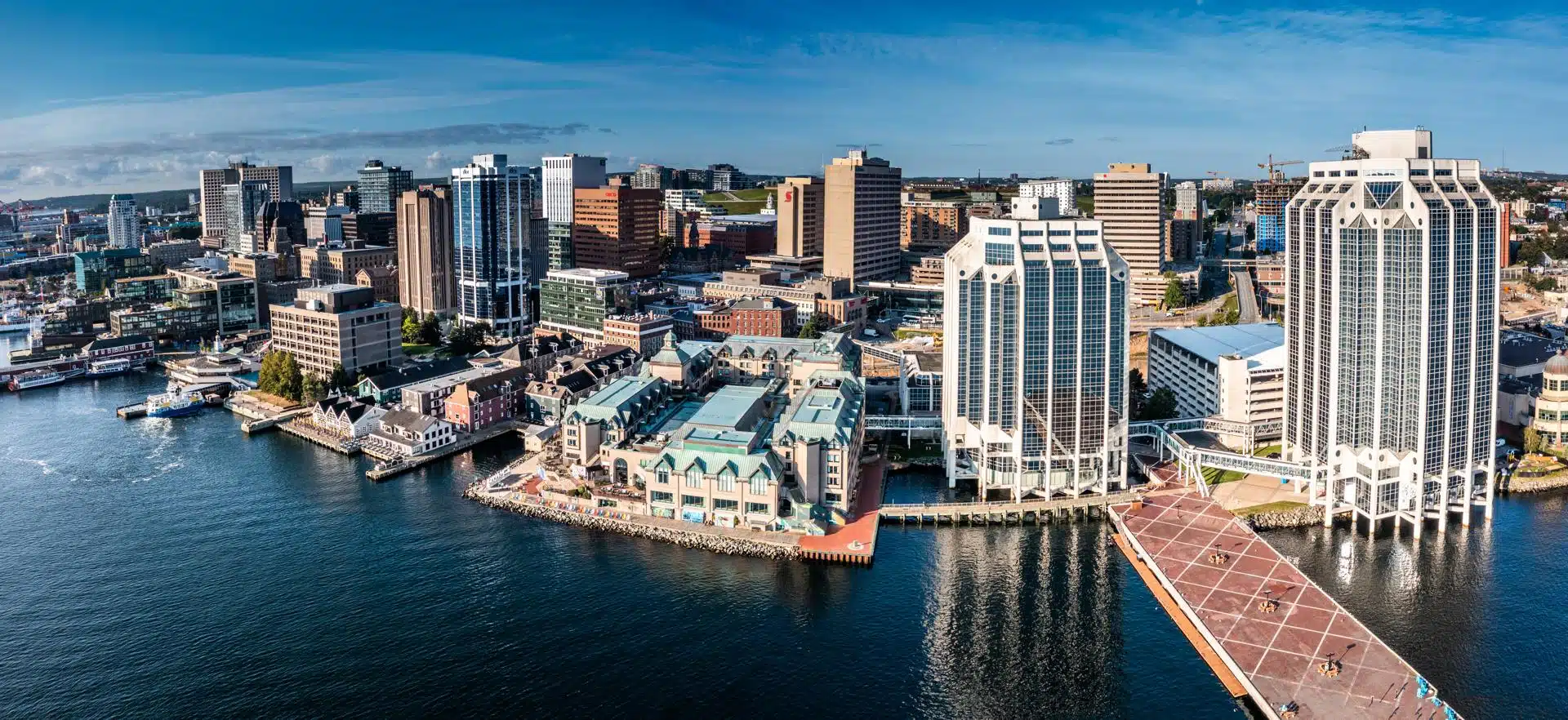 Panoramic aerial view of downtown Halifax, Nova Scotia, featuring a mix of modern office towers and historic waterfront buildings. The scene includes boardwalks, piers, and calm harbor waters under a clear blue sky.