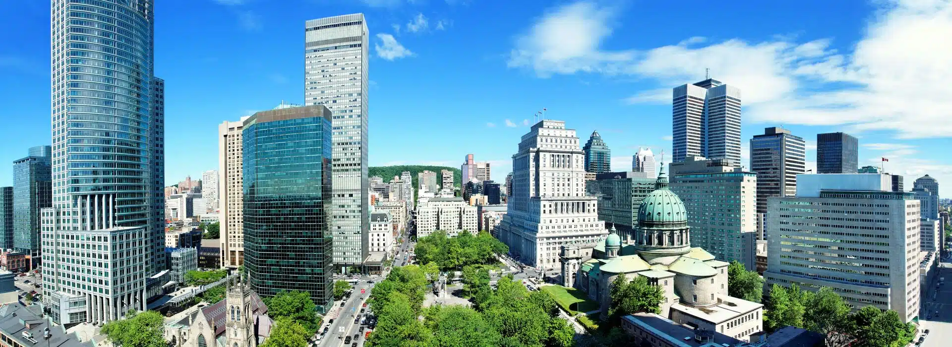 Panoramic view of downtown Montreal showcasing a mix of modern glass skyscrapers and historic architecture, including the green-domed Mary, Queen of the World Cathedral. Tree-lined boulevards and a bright blue sky complete the vibrant urban scene.