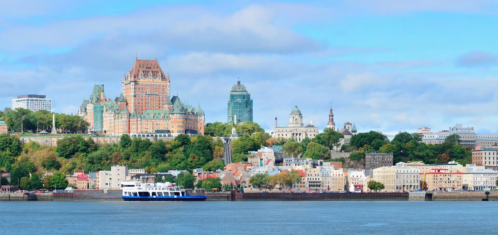 Scenic view of Old Quebec from across the St. Lawrence River, with the iconic Château Frontenac dominating the skyline. Historic buildings with colorful façades line the waterfront, framed by lush greenery and a ferry crossing the calm water.