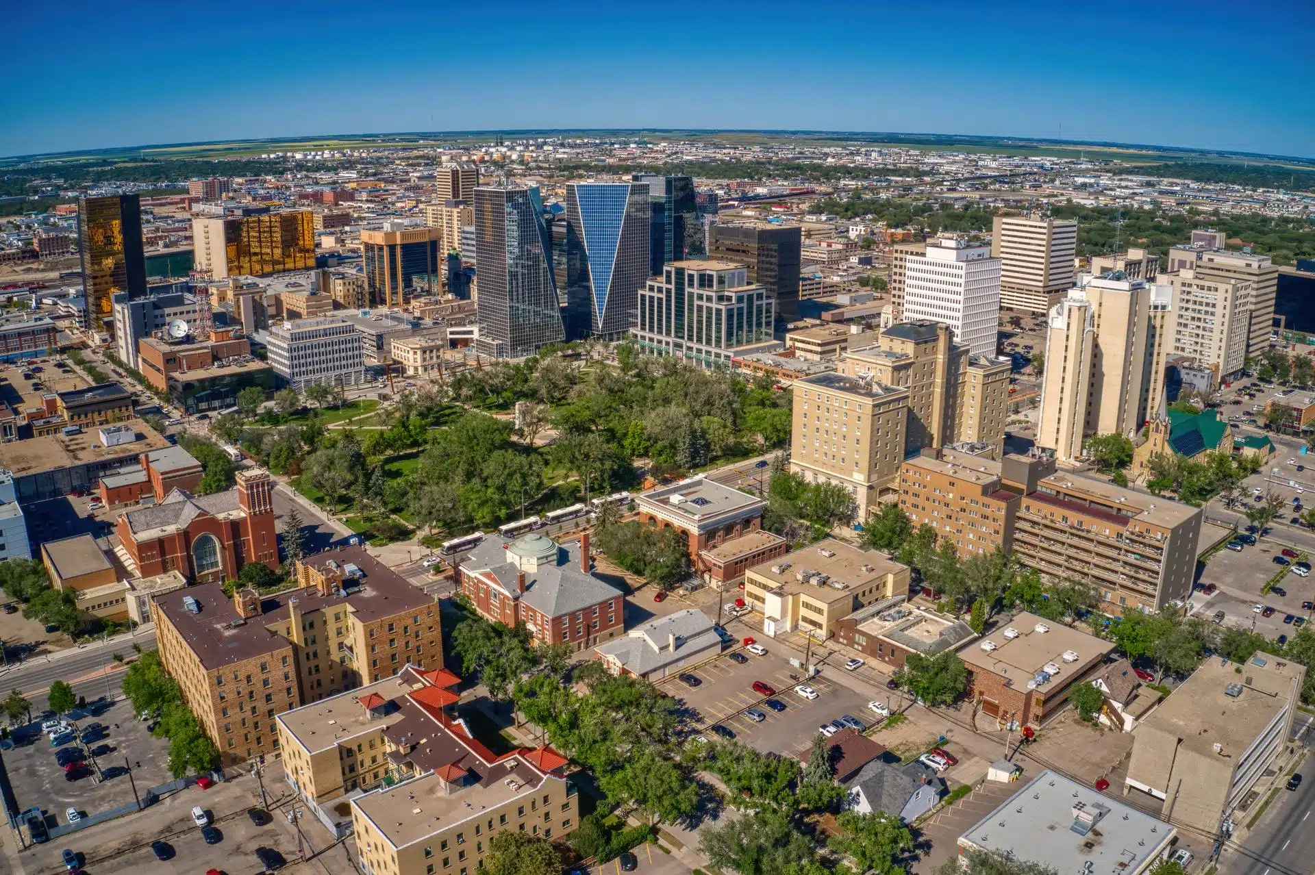 Aerial view of downtown Regina, Saskatchewan, featuring a mix of modern glass towers and historic buildings surrounding a central green space. The cityscape stretches into the horizon under a bright blue sky, highlighting urban development amid a prairie landscape.