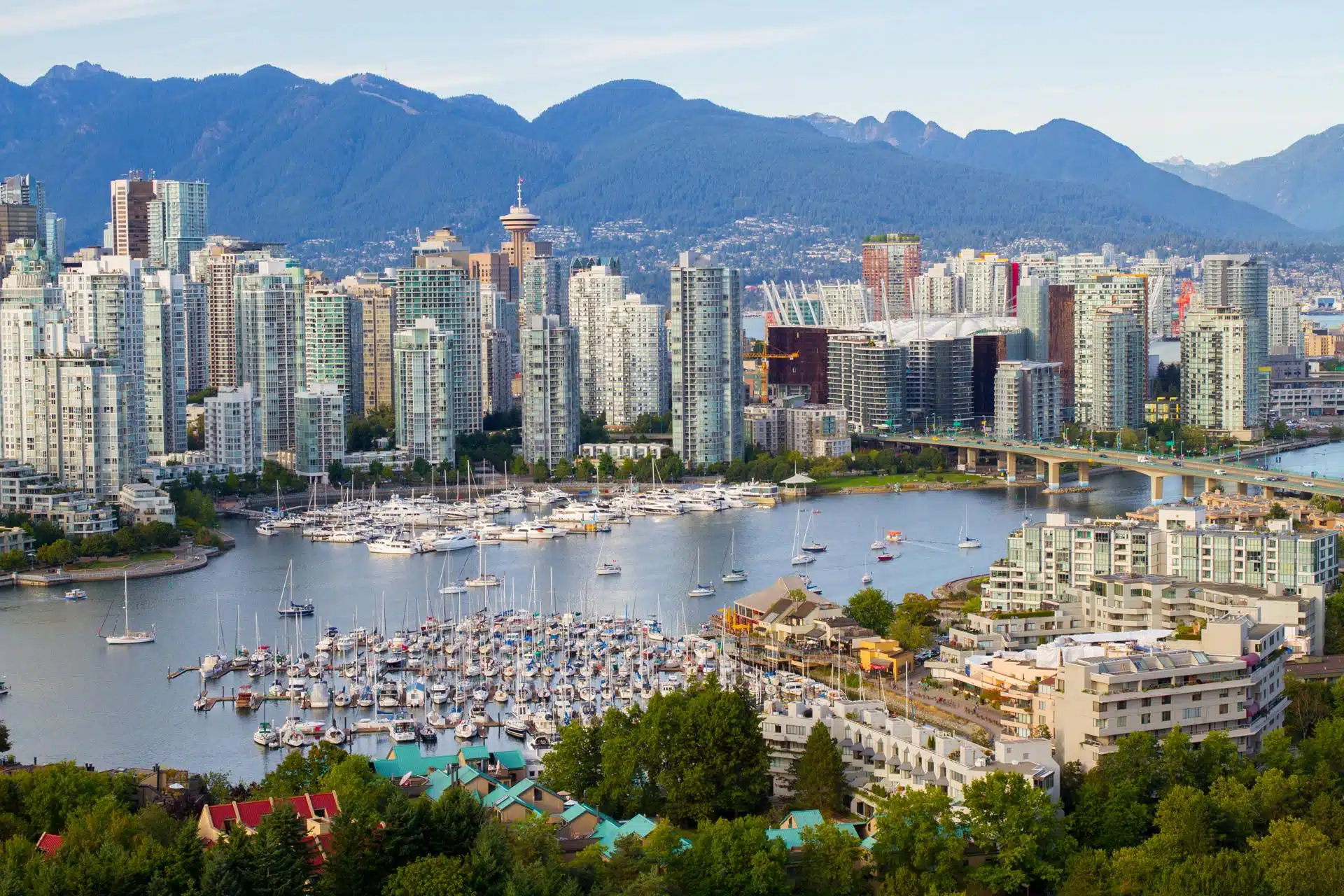 Wide-angle view of downtown Vancouver, British Columbia, showing a dense cluster of modern high-rise buildings along the waterfront with boats docked in the marina. In the background, the North Shore Mountains rise under a partly cloudy blue sky, framing the city with natural beauty.