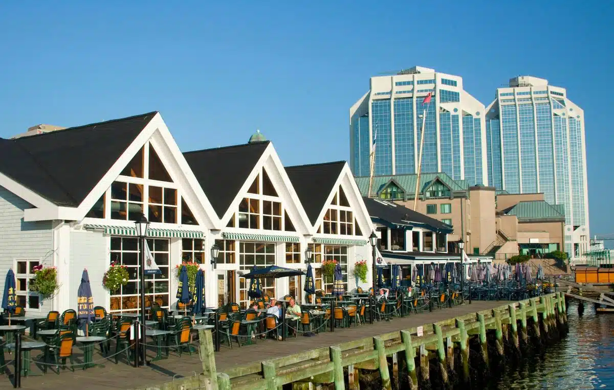 Scenic view of the Halifax waterfront showing a boardwalk lined with patio seating outside charming gabled buildings. In the background, tall glass office towers rise above the harbor on a sunny day.