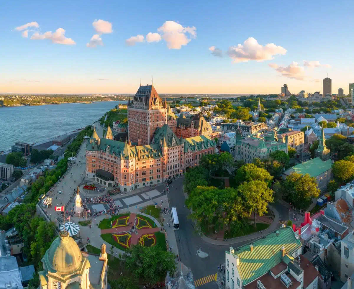 Aerial view of Old Quebec at sunset, highlighting the Château Frontenac and surrounding historic buildings with the St. Lawrence River stretching into the distance. The city is bathed in golden light, with tree-lined streets and a landscaped plaza filled with people.