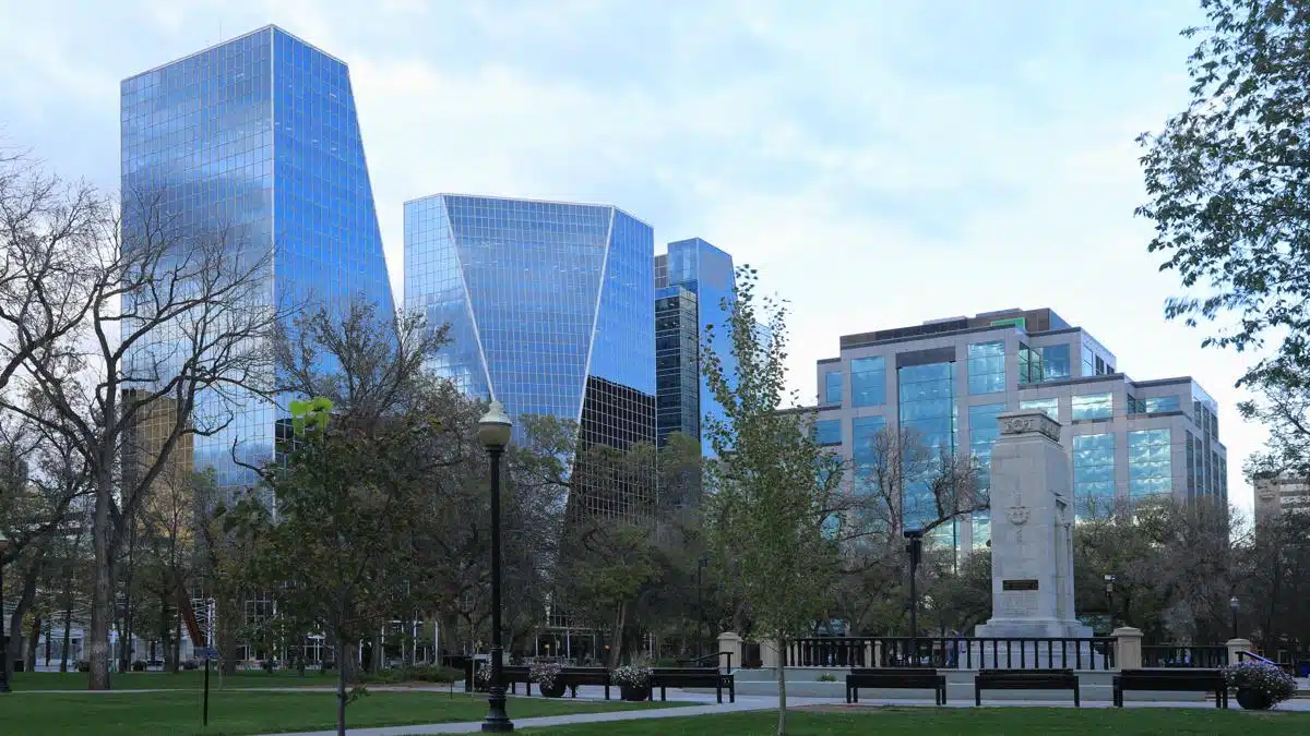 Downtown Regina skyline viewed from Victoria Park, with reflective glass towers and a war memorial monument in the foreground. Leafy trees and benches frame the urban scene on a calm, overcast day.