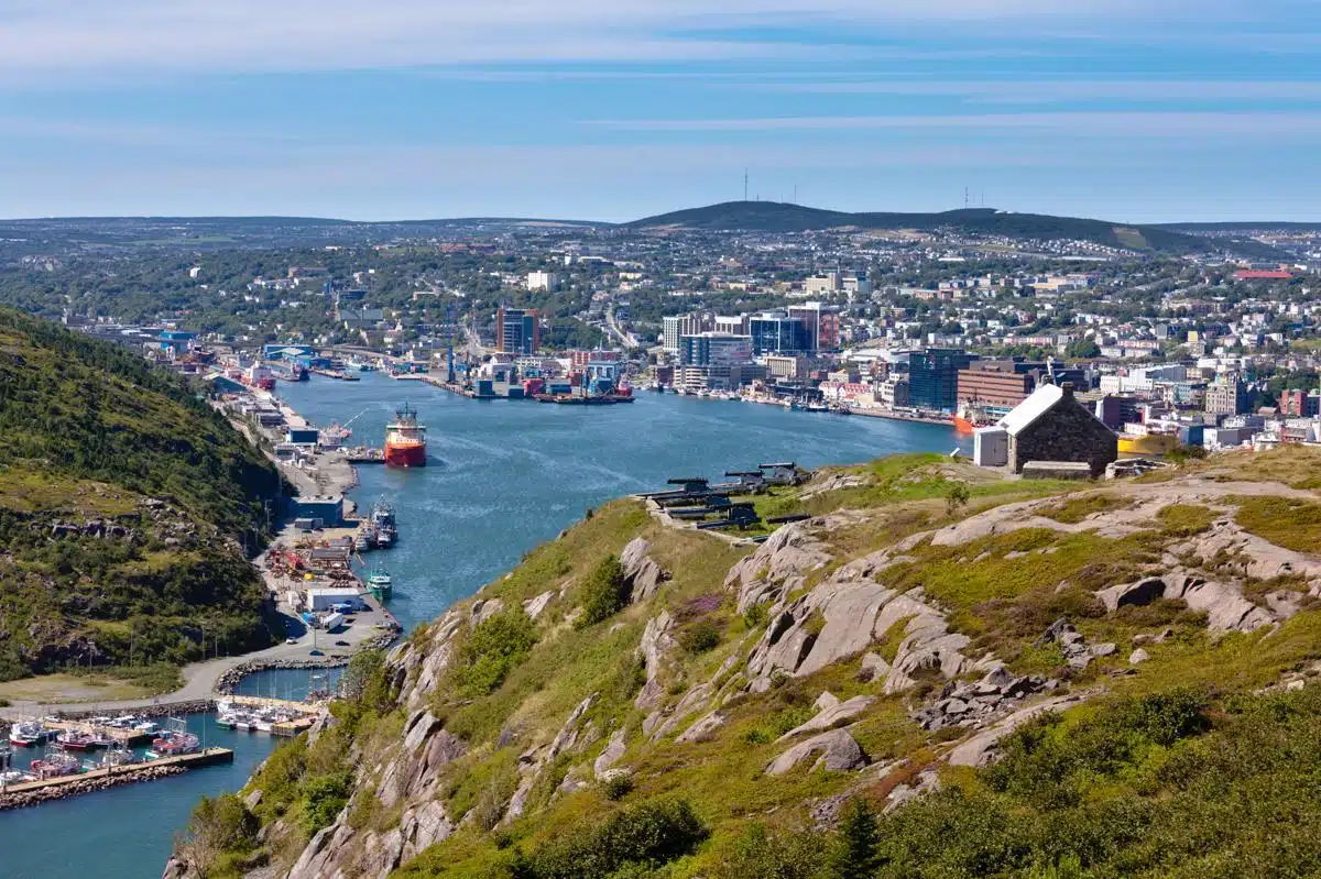 Scenic view overlooking the harbor and downtown St. John’s, Newfoundland, taken from Signal Hill. The image shows ships docked along the narrow inlet, modern buildings, and rolling hills under a bright blue sky.