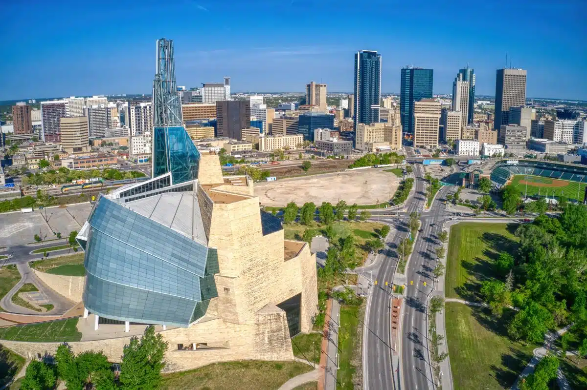 Closer aerial view of downtown Winnipeg showcasing the Canadian Museum for Human Rights with its iconic glass tower and curved stone design. Surrounding the museum are urban roads, green spaces, and the city skyline under a clear blue sky.