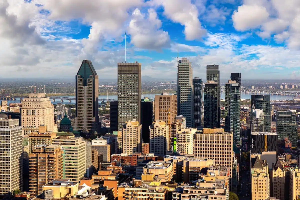 Aerial view of downtown Montreal with a mix of modern high-rises and older buildings, set against the backdrop of the St. Lawrence River and a long bridge. The skyline is under a partly cloudy sky, highlighting the city's urban density and architectural variety.